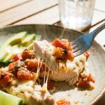 A close-up of a fork lifting a bite of cheesy salsa chicken with avocado on the plate.