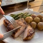 A close-up of a knife cutting into a juicy pork chop served with roasted vegetables.