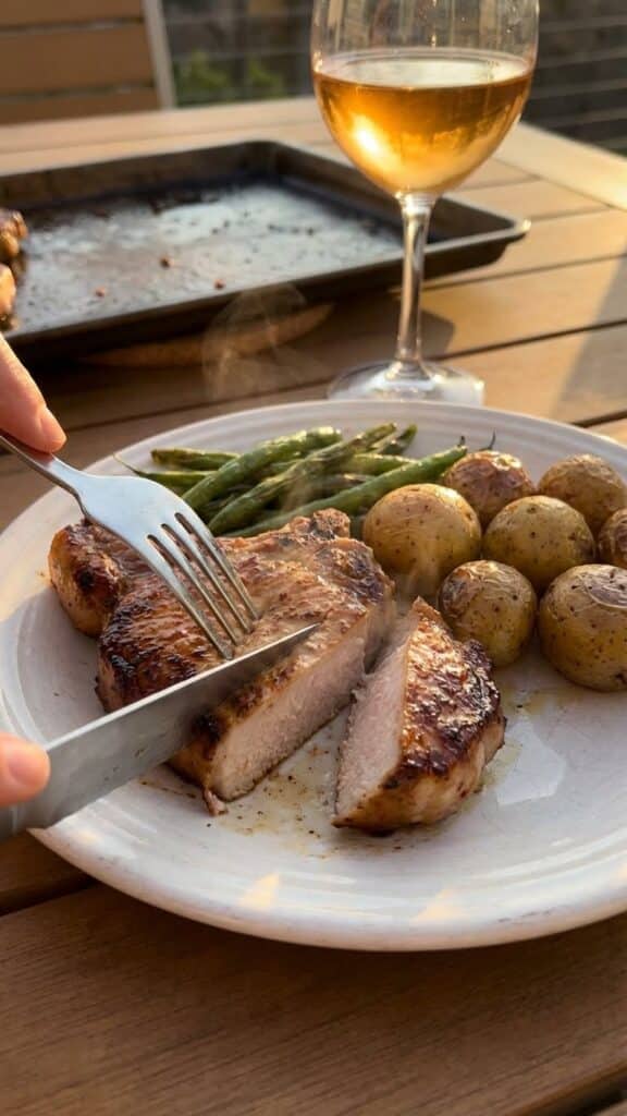 A close-up of a knife cutting into a juicy pork chop served with roasted vegetables.