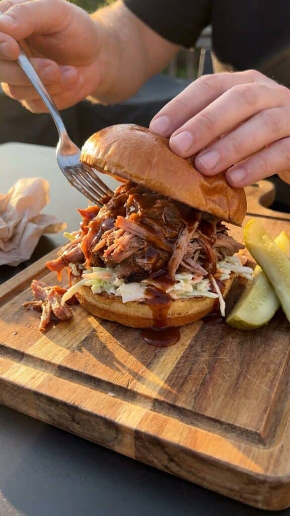 A close-up of a large BBQ pulled pork sandwich topped with coleslaw on a wooden board.