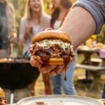 A close-up of a hand holding a large, messy BBQ pulled pork sandwich topped with coleslaw.