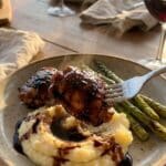 A close-up of a fork lifting a piece of balsamic glazed chicken over mashed potatoes and asparagus.
