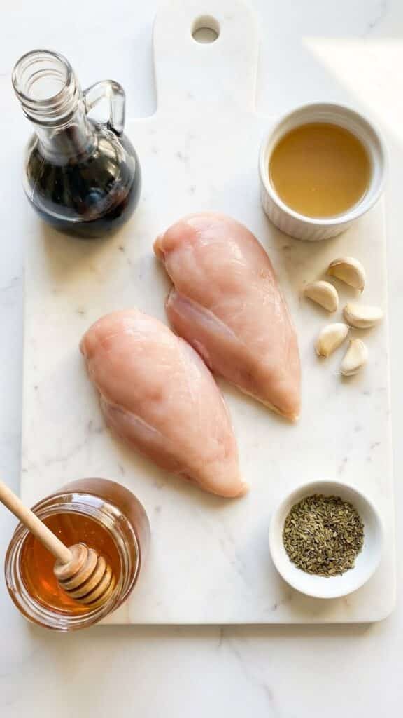 A flat lay showing raw chicken, balsamic vinegar, honey, garlic, and herbs on a marble board.