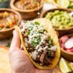 A close-up of a hand holding a loaded shredded beef taco with onion, cilantro, cheese, and crema, with a lime being squeezed over it.