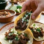 A close-up of a hand lifting an authentic street-style shredded beef taco topped with onion and cilantro from a plate.