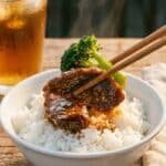 A close-up of chopsticks lifting glossy beef and broccoli from a bowl of white rice.