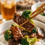 A close-up of chopsticks lifting a piece of glossy beef and broccoli from a bowl of white rice.