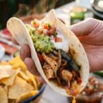 A close-up of a hand holding a flour tortilla loaded with chicken fajita filling, guacamole, and sour cream.