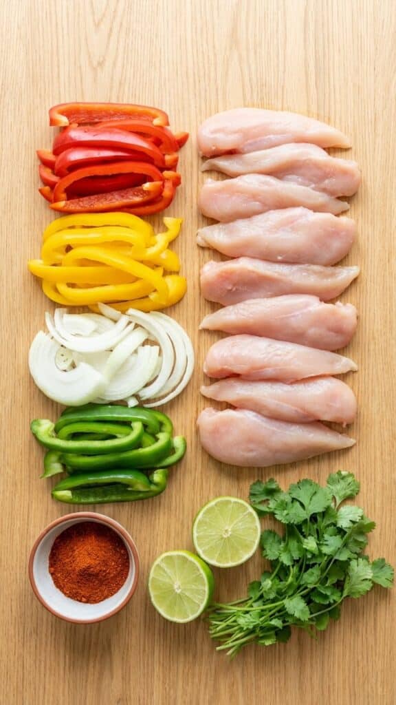A flat lay showing raw chicken strips, sliced red, green, and yellow peppers, onions, fajita seasoning, and limes.