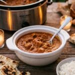 A close-up of a bowl of chicken tikka masala with a spoon, served with naan bread and rice on a wooden table.