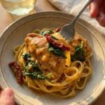 A close-up of a fork lifting a bite of creamy Tuscan chicken and spinach over pasta from a plate.