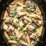 A top-down view inside a slow cooker filled with creamy chicken pasta with spinach and sun-dried tomatoes, topped with parmesan.