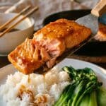 A close-up of a spatula lifting a piece of flaky, glazed salmon over rice and bok choy.