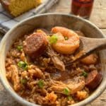 A close-up of a spoon lifting a bite of shrimp, sausage, and rice jambalaya from a bowl.