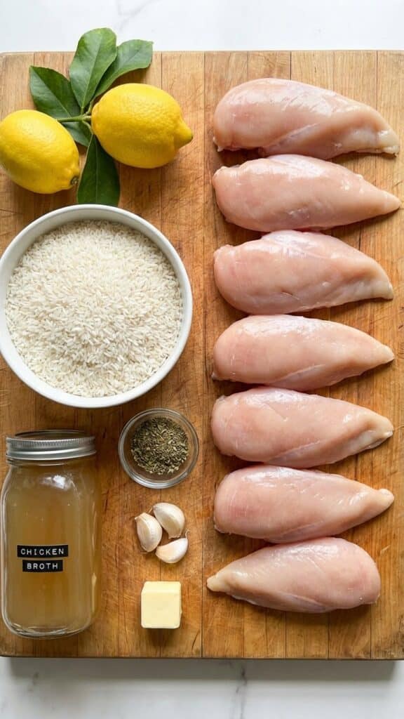 A flat lay showing raw chicken, uncooked rice, lemons, broth, and herbs on a wooden board.