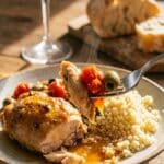 A close-up of a fork lifting a bite of Mediterranean chicken, olive, and tomato over a bed of couscous.