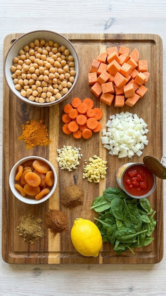 A flat lay showing raw sweet potatoes, chickpeas, carrots, spinach, apricots, and small piles of colorful spices like turmeric and cinnamon on a wooden board.