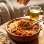A close-up of a spoon lifting sweet potato and chickpea stew from a bowl served over couscous with naan bread on the side.