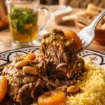 A close-up of a fork lifting a tender piece of lamb tagine with sauce and apricots from a bed of couscous.