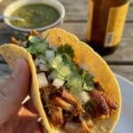 A close-up of a hand holding a corn tortilla taco filled with crispy pork carnitas, onion, and cilantro.