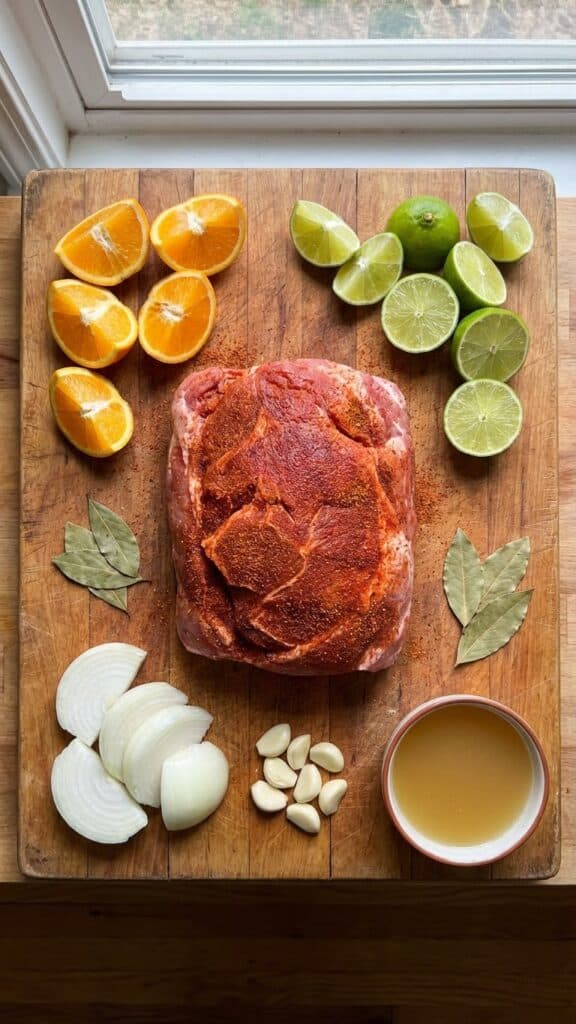 A flat lay showing a spice-rubbed raw pork shoulder, oranges, limes, onions, garlic, and broth on a wooden board.