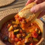 A close-up of a piece of crusty bread being dipped into a bowl of vegetable ratatouille.