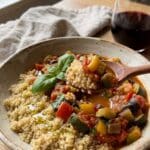 A close-up of a bowl with quinoa topped with ratatouille stew, with a spoon lifting a bite.