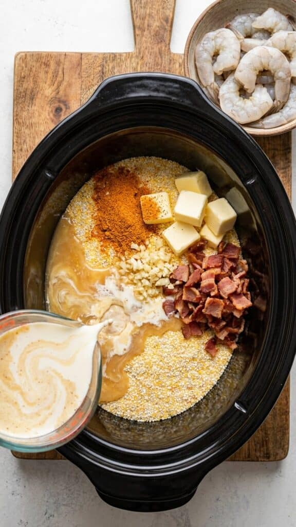 A flat lay showing grits, broth, cream, butter, and spices being added to a slow cooker, with a separate bowl of raw shrimp to the side.