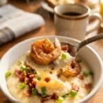A close-up of a spoon lifting a bite of creamy grits and a Cajun shrimp from a bowl.