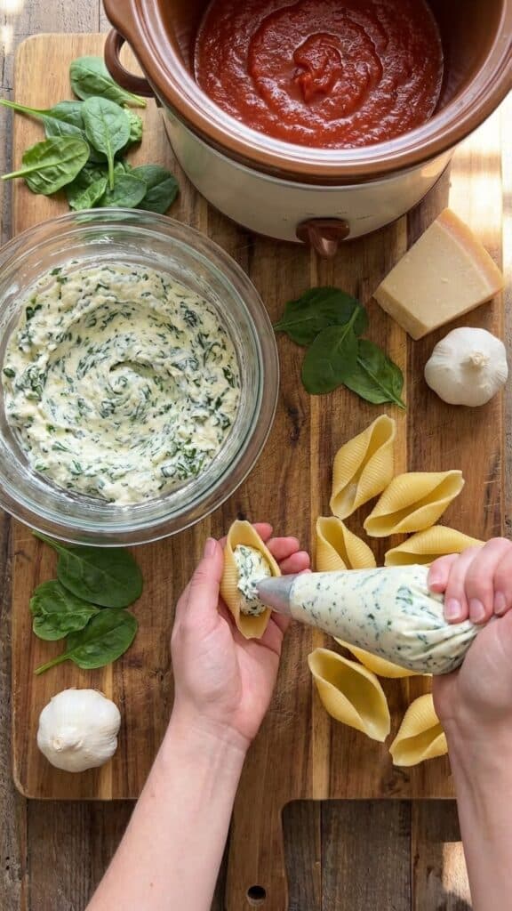 A flat lay showing the process of filling jumbo pasta shells with a spinach and ricotta mixture, next to a slow cooker pot containing marinara sauce.