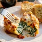 A close-up of a fork cutting into a cooked stuffed shell on a plate, revealing the creamy spinach and ricotta filling.