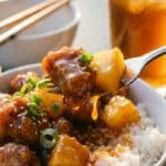 A close-up of a fork lifting a piece of glossy sweet and sour pork and pineapple over a bowl of rice.