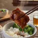 A close-up of chopsticks lifting a piece of glossy teriyaki chicken from a bowl of rice and broccoli.