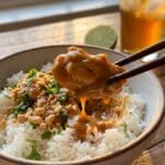 A close-up of chopsticks lifting a piece of saucy peanut chicken from a rice bowl.