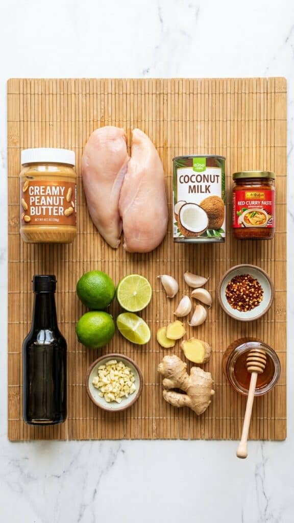 A flat lay showing chicken, peanut butter, coconut milk, curry paste, soy sauce, lime, and ginger on a bamboo mat.