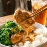A close-up of chopsticks lifting a piece of saucy peanut chicken from a bowl of rice and broccoli.