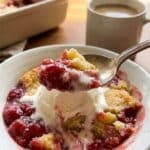 A close-up of a bowl containing a scoop of strawberry cheesecake dump cake topped with melting vanilla ice cream, with a spoon lifting a bite.