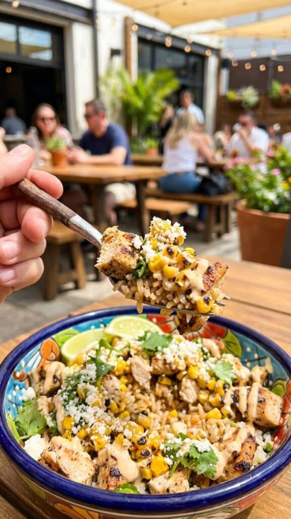 A close-up POV shot of a fork lifting a mixed bite of chicken, rice, and creamy street corn from a bowl.