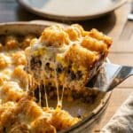 A close-up of a spatula lifting a square of tater tot casserole, showing the beef layer and cheese pull.