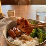 A close-up of chopsticks lifting a piece of glossy teriyaki chicken from a rice bowl with broccoli.