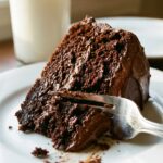 A close-up of a fork cutting into a moist slice of chocolate cake with fudge frosting.