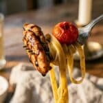 A close-up of a fork lifting balsamic chicken, tomato, and pasta.