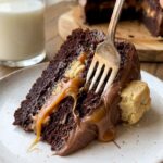 A close-up of a fork cutting into a slice of chocolate cake with caramel oozing out and cookie crumbs visible.