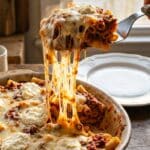 A close-up of a spatula lifting a serving of baked ziti with a long, gooey cheese pull.