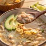 A close-up of a spoon lifting a bite of creamy white chicken chili with avocado and tortilla chips from a bowl.