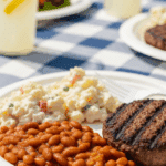 A generous serving of 3-bean baked beans on a BBQ plate next to a burger and potato salad, set in a sunny outdoor picnic atmosphere.