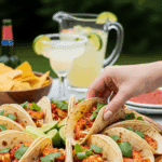 A party platter of spicy shrimp tacos with a hand reaching for one, with margaritas in the background.