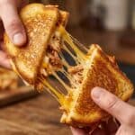 Hands pulling apart a BBQ chicken grilled cheese sandwich showing a long stretchy cheese pull.