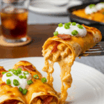 Plated Bacon-Ranch Chicken Enchiladas with filling showing, served with sour cream on a dinner table.