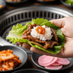A hand making a lettuce wrap with beef bulgogi, rice, and ssamjang, set against a background of various Korean side dishes (banchan).